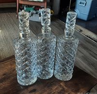 Three vintage crystal liquor decanters with diamond-cut pattern and matching stoppers displayed on wooden floor.