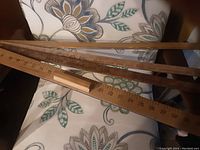 Four vintage wooden yardsticks placed on and beside a patterned chair, showing full length and measurement markings, plus advertising text on some sticks.