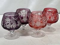 Four Ajka Marsala cut to clear crystal brandy glass snifters, two purple and two cranberry, arranged in two rows.
