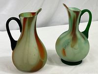 Both hand blown art glass pitchers shown side by side on a white background, displaying their complementary marbled colors and black handles.