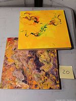 Two square abstract paintings stacked, one yellow with green and black swirls, and one purple and orange textured design, on a table