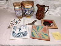 View of entire lot showing two ceramic drums, copper glaze pitcher, and five tile trivets with various decorations.