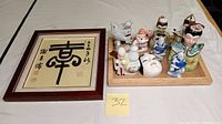 Overall view of framed Asian artwork and porcelain figurines displayed on a wooden tray, tray not included in lot.