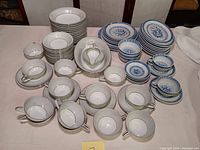 Wide view showing stacked Noritake Sabrina teacups and saucers with white and silver floral pattern along with stacks of Asian blue and white dragon themed plates and bowls.
