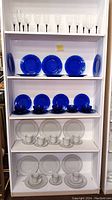 Full shelf showing white china with plates, cups, saucers on lower shelves, cobalt blue glass dinnerware and bowls on middle shelves, clear glasses with black stems on top shelf.