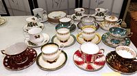 Overview of 12 different vintage decorative china teacups with matching saucers arranged on a table showing various colors and designs including Christmas and floral motifs.