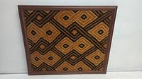Framed Kuba textile fabric with intricate dark brown and tan geometric patterns, displayed on a white surface.