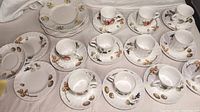 Wide overhead view of 9 Mikasa Maxima teacups and saucers with floral patterns, and 5 Royal Caldon Bristol ironstone luncheon plates arranged on a white cloth.