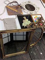Photo showing two large ornate mirrors, one rectangular with detailed ornate edges and one oval with floral scroll design. White round enameled floral metal mirror also visible on table along with two purses and three framed artworks.
