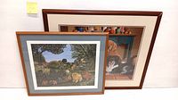 Both lithographs framed, Kip Kopatch lithograph in front showing a colorful outdoor dog scene, Braldt Bralds lithograph behind with dog and pottery.