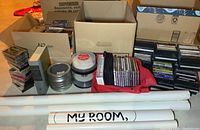 Box with multiple stacks of cassette tapes, several DVDs, blank DVD/CD spindles, and rolled posters visible on a table.