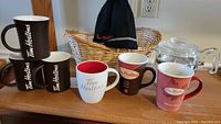 Six Tim Hortons coffee mugs arranged with a black Tim Hortons toque and wicker basket, alongside a clear glass coffee percolator.