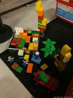 Duplo blocks, figures, palm tree, and wheeled base with blocks in silver box, showing colorful blocks and assembled pieces on floor.
