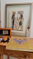 Framed Egyptian papyrus art showing two figures with hieroglyphs, wooden scarab box, Egyptian bookmark, shadowbox, and small matching framed art on a wooden surface.