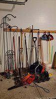 Photo showing three black metal shepherd's hooks, two black metal trellises, several long-handled gardening tools including cultivator, hoe, shovel, and spade, coiled extension cords, leaf blower, and various small hand tools.