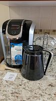 Keurig 2.0 coffee maker, black coffee pot, metal pod holder placed on countertop with coffee descaling powder box and measuring scoop