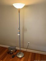 Full view of metal floor lamp turned on showing the frosted upturned shade and goose neck task light near the middle, Bose SoundDock, power cords, and remote on the floor base nearby