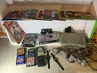 PlayStation console with controllers, Atari cartridges, magazines, books, boxed D-Link adapter, electronic game and action figure visible