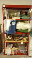 Front view of red metal shelving unit loaded with various household items including blue storage bin, fabric, and craft supplies.