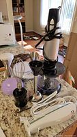 Hamilton Beach immersion blender with attached cord, whisk, blending shaft, and chopping bowl on a tabletop with natural light.