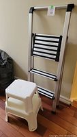 Folding aluminum step ladder leaning against wall with two white plastic step stools stacked in front