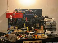Various hand tools arranged on a workbench, including screwdrivers, clamps, hand saw, electric drill and saw, red metal toolbox on pegboard, and small plastic drawers with hardware items