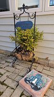 Front view of metal planter with live plant and hanging WELCOME sign on paved surface against house siding. Decorative concrete welcome stone with frog design placed next to planter.