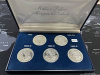 Full view of five silver-plated replica Morgan silver dollars arranged in a blue velvet-lined case with inscriptions showing years and mint marks, inside a hinged presentation box with satin lining.
