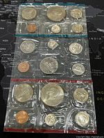 Photo of three sets of coins sealed in original packaging, showing various denominations including pennies, nickels, dimes, quarters, half-dollars, and dollar coins, from both US and Canada in proof-like condition.