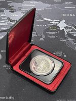 Round silver coin in protective capsule inside red velvet-lined black display box, showing coin and presentation case.