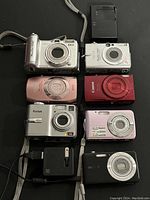 Eight cameras and one charger laid out front-side up on black background