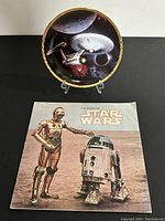 Plate displayed on a clear stand with booklet in front, showing a Star Trek space theme plate and a Star Wars booklet cover.