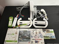 Photo showing Nintendo Wii console in vertical stand with cord, 2 white Wii Remote controllers, 2 white Nunchuck controllers, 2 white Wii Wheel attachments, and 4 game cases visible laid out beneath.