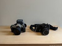 Two vintage cameras positioned side-by-side, Canon AE-1 on the left with silver/black body, GAF L-ES/2 on the right with black body and lens cap on.