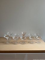 Ten assorted champagne coupe glasses displayed on a wooden surface, showing variety in style and size, including one pink tinted glass.