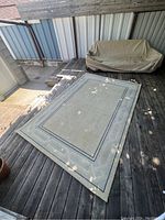 Full view of cream-colored rectangular rug laid on wooden floor outdoors, showing geometric border pattern and two parallel lines framing the inner area.