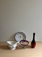 Photo of four vintage homegoods: a blue-trimmed plate, a white floral relief gravy boat, a berry-colored glass bowl, and a red-black ombre glass vase on a wooden surface against a plain wall.