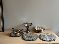 Photo showing all silver items on a wood surface including two trivets, serving dish with glass, vase, and decorative boat.