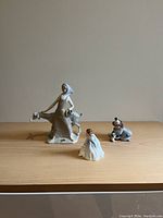 Three figurines displayed on a wooden surface against a beige wall: woman with goat, clown, and young girl.