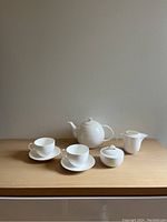 White ceramic tea set arranged on wooden surface against plain wall, includes teapot, creamer, sugar bowl with lid, and 2 cups with saucers.