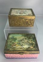 Two antique European style trinket boxes shown on clear stands. Top box is brass with etched designs and a floral tapestry lid. Bottom box is German porcelain with pink embossed base, pastoral scene on top, and brass trim.
