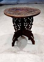 Front angle view of the antique Indian wooden folding round side table on a concrete floor, showing carved openwork legs and round tabletop.