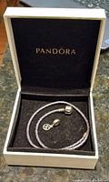 Gray braided leather double-wrap Pandora bracelet with silver clasp and charm pendant displayed inside black velvet-lined white Pandora box.