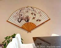 Large Chinese fabric wall fan mounted on wall with visible floral and bird painting, bamboo frame, above white dining chairs and a table.