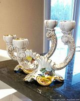 Front view of one ornate ceramic candlestick holder featuring two candle sockets with gilded trim, curved arms with gold detail, and a base painted with a small house and greenery scene.