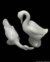 Two white porcelain goose figurines, one with neck extended and one with curved neck, both on base