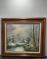 Full frontal image of framed winter landscape painting showing scene with cabin, snow, trees and water body.