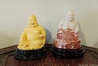 Two Laughing Buddha figurines on pedestals, one yellow resin and one white/red marble, shown together on a wooden surface.