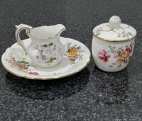 Three porcelain pieces set on dark granite countertop: a creamer on saucer, a lidded jam jar, and a separate saucer, each decorated with floral patterns and gold trim.