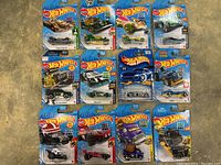 12 Hot Wheels cars in original packaging laid out on carpet, showing the variety of sizes and styles of die-cast vehicles in blue blister cards.